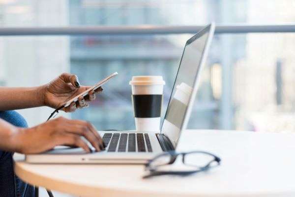 A laptop open with person working with smart phone plugged in. All set against a glass backdrop with coffee cup in distance.