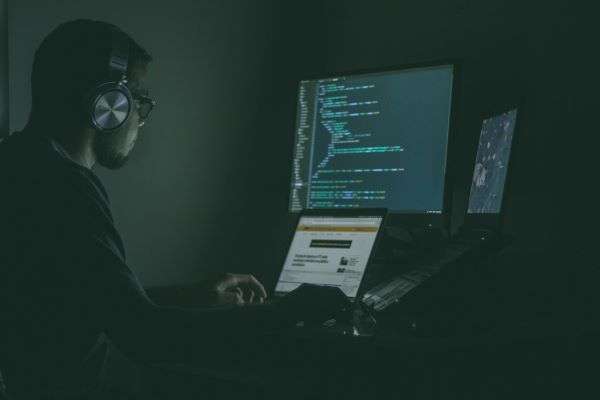 A computer hacker working across a laptop and larger screen with light off.